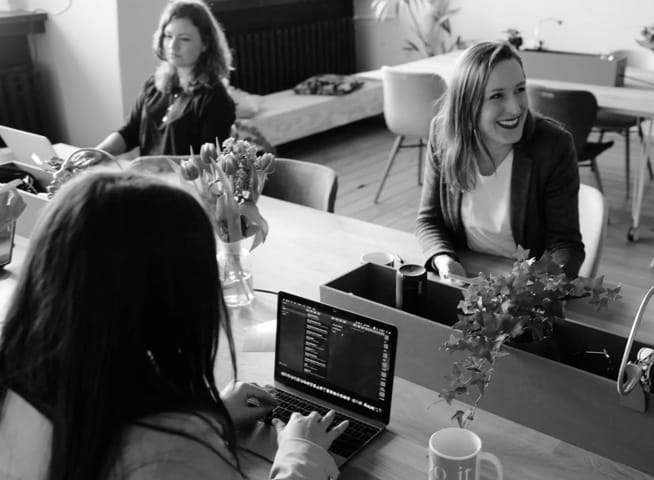 A black and white photo of a group of ladies working together on a table in front of their laptops, laughing and having fun.